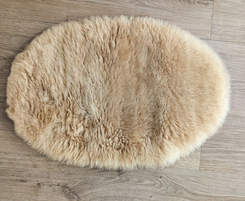 May include: A light brown sheepskin rug with a soft, fluffy texture. The rug is oval shaped and is laying flat on a wooden floor.