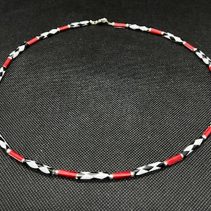 Könnte beinhalten: Halskette mit abwechselnd schwarzen und weißen rautenförmigen Perlen und roten zylindrischen Perlen. Die Kette wird durch silberfarbene Elemente verbunden. Der Schmuck wird vor einem dunklen Hintergrund präsentiert und hat einen Silberverschluss.
