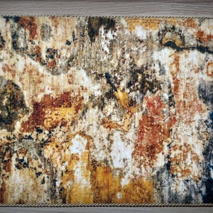 May include: Rectangular rug with an abstract design in shades of brown, yellow, and blue. The rug has a textured appearance with a rope-like border. The number "3" is visible in the upper left corner.