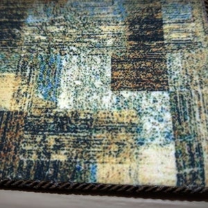 May include: Close-up of a rectangular rug with an abstract pattern in shades of brown, beige, blue, and green. The rug has a textured appearance and a dark brown rope border. The design features a patchwork-like arrangement.
