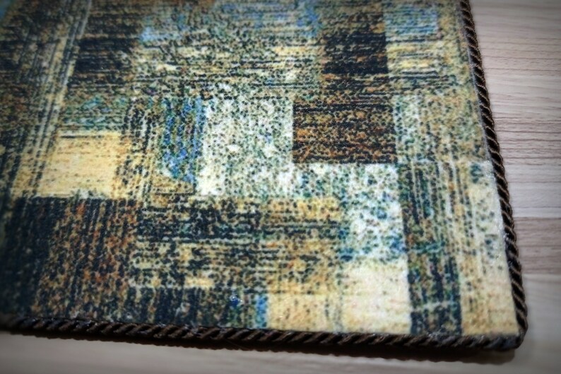 May include: Close-up of a rectangular rug with an abstract pattern in shades of brown, beige, blue, and green. The rug has a textured appearance and a dark brown rope border. The design features a patchwork-like arrangement.