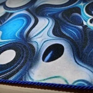 Peut inclure: Gros plan sur un tapis &agrave; motifs bleus et blancs avec une bordure en corde bleu fonc&eacute;. Le motif pr&eacute;sente des motifs tourbillonnants dans les tons de bleu, de blanc et de noir, cr&eacute;ant un aspect fluide et abstrait. Les bords du tapis sont soigneusement finis.