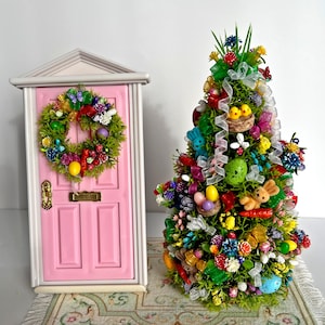 Op de afbeelding: Een roze miniatuurdeur met een krans versierd met kleurrijke paaseieren en bloemen, naast een miniatuur paasboom versierd met kleurrijke eieren, bloemen, linten en een konijn.
