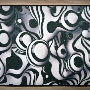 Peut inclure: Un tapis rectangulaire avec une bordure en corde noire. Le tapis pr&eacute;sente un motif abstrait noir, blanc et gris avec des formes sph&eacute;riques et ondul&eacute;es. Le design cr&eacute;e un effet 3D, avec les formes semblant flotter.