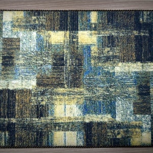 May include: Rectangular placemat with a textured, abstract design in shades of blue, brown, and yellow. The edges are trimmed with a dark brown rope-like border. The design features a grid-like pattern.