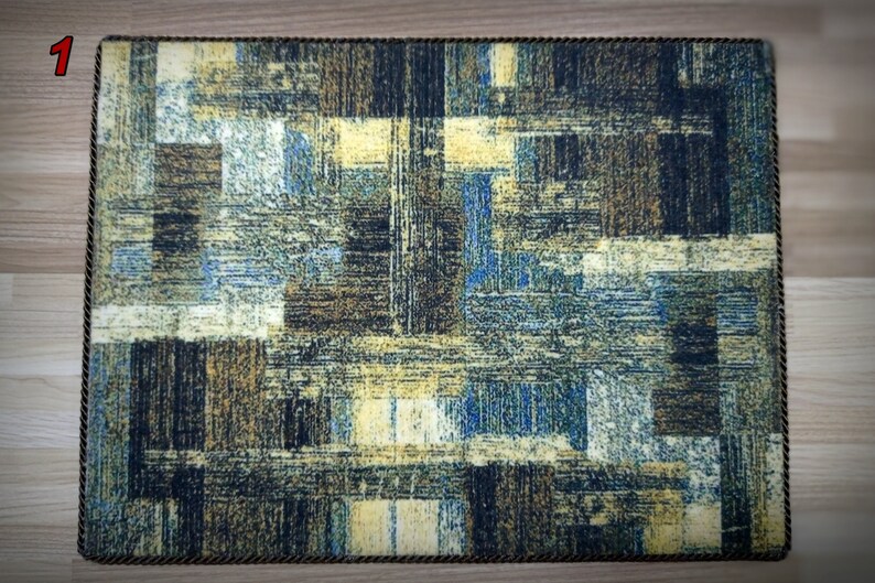May include: Rectangular placemat with a textured, abstract design in shades of blue, brown, and yellow. The edges are trimmed with a dark brown rope-like border. The design features a grid-like pattern.