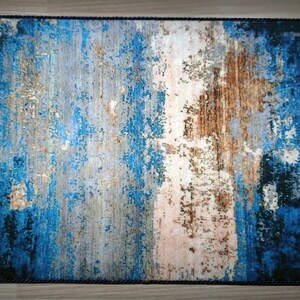 May include: Rectangular rug with a distressed abstract design in shades of blue, brown, and gray. The rug has a dark blue rope border, adding a textured edge to the overall aesthetic. The design gives the rug a vintage or weathered appearance.