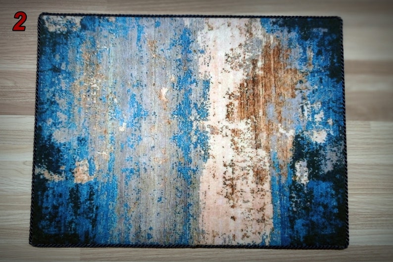 May include: Rectangular rug with a distressed abstract design in shades of blue, brown, and gray. The rug has a dark blue rope border, adding a textured edge to the overall aesthetic. The design gives the rug a vintage or weathered appearance.