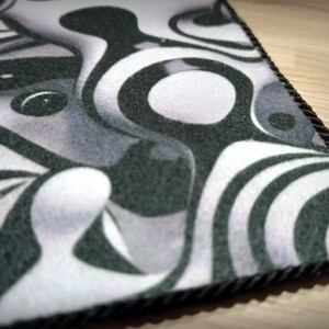 Peut inclure: Un tapis d&eacute;coratif avec un motif abstrait noir, blanc et gris. Le motif pr&eacute;sente des formes tourbillonnantes et des &eacute;l&eacute;ments circulaires. Le tapis a une bordure en corde noire et est probablement fait d'un mat&eacute;riau doux semblable &agrave; du feutre.