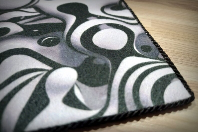 Peut inclure: Un tapis d&eacute;coratif avec un motif abstrait noir, blanc et gris. Le motif pr&eacute;sente des formes tourbillonnantes et des &eacute;l&eacute;ments circulaires. Le tapis a une bordure en corde noire et est probablement fait d'un mat&eacute;riau doux semblable &agrave; du feutre.