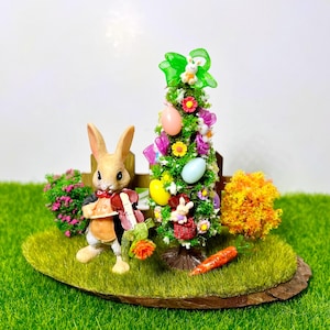 Op de afbeelding: Een miniatuur Pasen scène met een konijn dat een muziekinstrument vasthoudt, een gedecoreerde paasboom met kleurrijke eieren en linten, en een kleine struik met groene en gele bladeren. De scène is geplaatst op een bruine houten basis met groen gras.