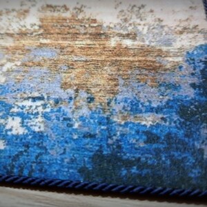 May include: Close-up of a rectangular rug with an abstract design. The rug features a blend of blue, brown, and white hues, with a textured appearance. A dark blue rope trim borders the rug's edge, adding a decorative element.
