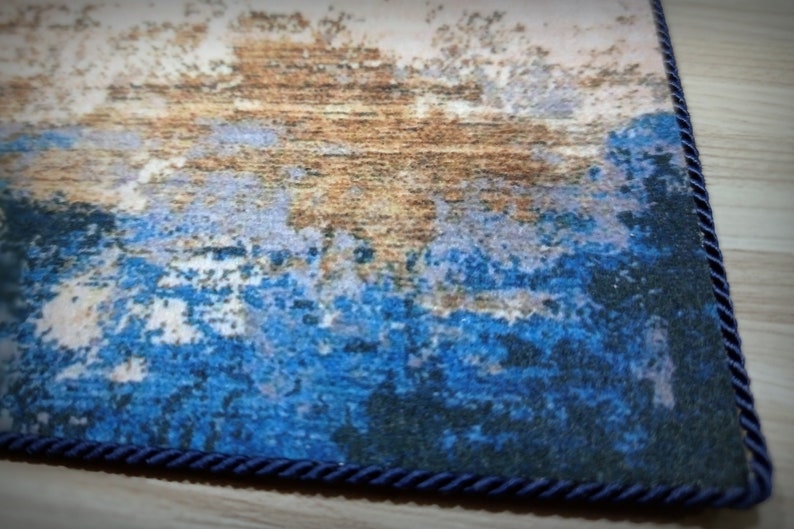 May include: Close-up of a rectangular rug with an abstract design. The rug features a blend of blue, brown, and white hues, with a textured appearance. A dark blue rope trim borders the rug's edge, adding a decorative element.