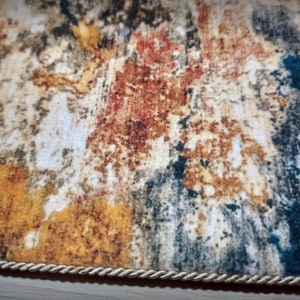 May include: A rectangular rug with an abstract design in shades of yellow, brown, red, and blue. The rug has a textured appearance and is bordered with a cream-colored rope trim. The rug's design resembles a watercolor painting.