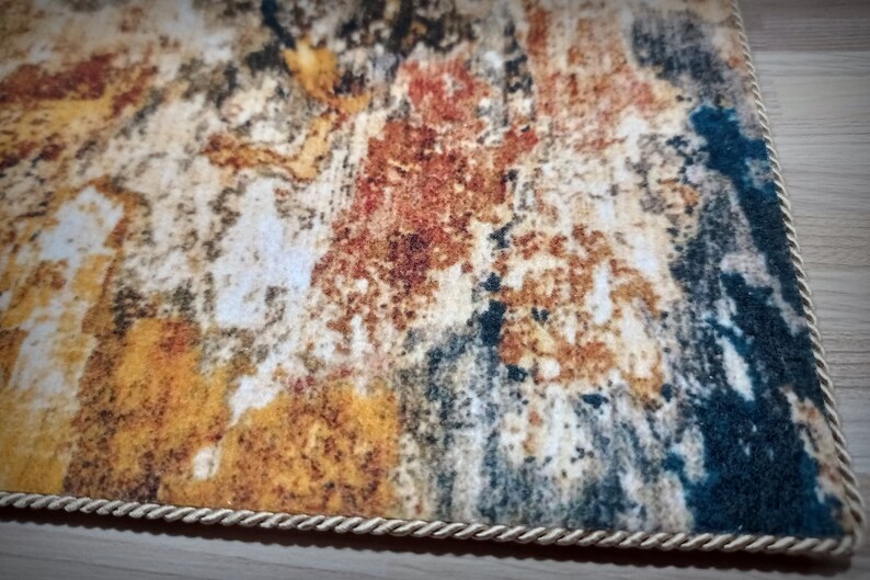 May include: A rectangular rug with an abstract design in shades of yellow, brown, red, and blue. The rug has a textured appearance and is bordered with a cream-colored rope trim. The rug's design resembles a watercolor painting.
