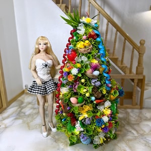 Op de afbeelding: Een miniatuur paasboom versierd met kleurrijke eieren, bloemen, kuikens en linten. De boom is ongeveer 30 cm hoog en gemaakt van groen vilt en andere materialen. Een pop staat naast de boom.