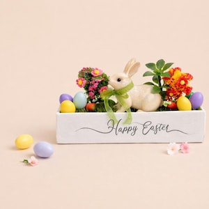 May include: A white rectangular planter box with a bunny, colorful eggs, and flowers. The bunny has a green ribbon. The box has the words "Happy Easter" in script. Easter decor.