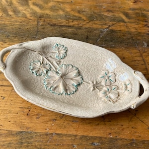 Può includere: Un piatto beige in ceramica a forma di foglia con superficie strutturata. Il piatto presenta un motivo floreale in rilievo con accenti verdi e bianchi. Ha un manico ad ogni estremità ed è appoggiato su una superficie di legno.