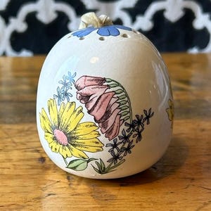 Op de afbeelding: Een witte, eivormige zoutstrooier van keramiek met een bloemenontwerp. De strooier heeft een gele madelief, roze en groene bloemen en blauwe accenten. De bovenkant heeft kleine gaatjes om zout te doseren en een beige touwtje.