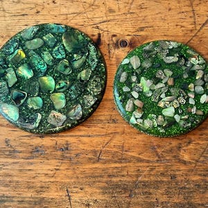 Pode incluir: Duas bases redondas com base verde escuro, incrustadas com pedras verdes e cinzentas iridescentes. As bases estão sobre uma superfície de madeira, mostrando seu design único e natural. Perfeitas para adicionar um toque de decoração natural.