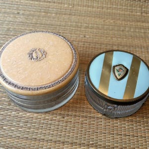 Peut inclure: Deux poudriers anciens. L'un est rond avec un couvercle beige clair, une bordure décorative et un monogramme central. L'autre est ovale avec un couvercle à rayures bleu clair et or, et un emblème floral. Les deux ont des bases en verre.