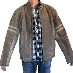 Op de afbeelding: Een bruin suède jasje met een ritssluiting en twee lichtbruine strepen op elke mouw. Het jasje wordt gedragen over een zwart-wit geruit overhemd en een blauwe spijkerbroek.