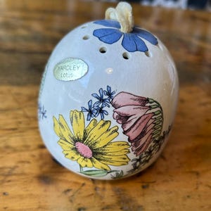 May include: A white, egg-shaped Yardley Lotus ceramic container with floral designs in yellow, pink, and blue. The container has small holes on top and a gold ribbon loop. The Yardley Lotus label is visible on the side.