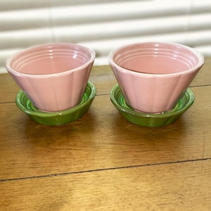 May include: Two matching ceramic bowls in a soft pastel pink color, each featuring a fluted exterior design. The bowls are each set inside a matching green ceramic saucer that also features a fluted edge, creating a coordinated set of two.
