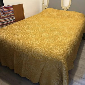 May include: A mustard yellow bedspread with a repeating floral pattern covers a bed. The headboard is grey with a white decorative trim. A colourful blanket is visible on the left side of the bed.
