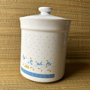 Op de afbeelding: Witte keramische voorraadpot met deksel, versierd met blauwe stippen en een rij cartoon eenden met blauwe sjaals. De pot heeft een blauwe stippenband en een klein handvat op het deksel.