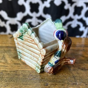 May include: A ceramic planter shaped like a log cabin with a glossy finish. The structure features light brown log-style walls and a green roof. Two small, colorful birds with blue, green, and yellow plumage are perched on a brown branch extension.