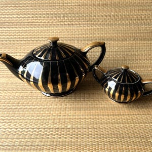 Può includere: Un set di teiera e lattiera in ceramica nera e oro. La teiera ha un beccuccio e un manico curvi, con dettagli dorati. La lattiera è una versione più piccola della teiera. Entrambi i pezzi presentano strisce dorate verticali.