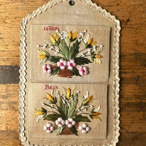 May include: A beige woven wall-hanging organiser with two pockets. Each pocket features embroidered floral arrangements in yellow, white, and pink with green leaves. The top pocket is labelled "LETTERS" and the bottom pocket is labelled "BILLS".