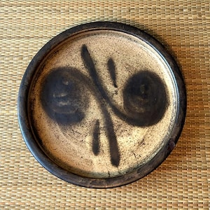 May include: A round ceramic plate with a dark brown rim and a beige center. The center features a dark brown abstract design with two spiral shapes and vertical lines. The plate has a rustic, handmade appearance.