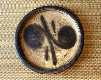 Op de afbeelding: Een ronde keramische plaat met een donkerbruine rand en een beige midden. Het midden heeft een donkerbruin abstract ontwerp met twee spiraalvormen en verticale lijnen. De plaat heeft een rustieke, handgemaakte uitstraling.