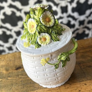 May include: A white ceramic jar with a woven basket texture and a lid featuring a sculpted cluster of yellow daisies with green leaves. A green vine handle extends from the side of the jar, and a small yellow flower is attached to the front surface.