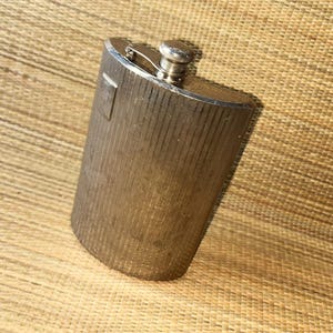 以下が含まれることがあります： 縦縞模様のテクスチャが施された銀色のフラスコ。 フラスコは、ネジ式のキャップが付いた丸みを帯びた上部と、前面の小さな長方形のディテールを備えています。 フラスコは金属製です。