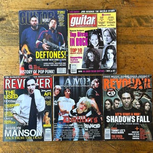Könnte beinhalten: Eine Sammlung von sechs Musikmagazinen, darunter "Guitar World" und "Revolver", mit Rock- und Alternative-Musik-Künstlern. Die Cover zeigen Bands wie Deftones, Marilyn Manson und The Distillers. Die Zeitschriften liegen auf einer Holzoberfläche.