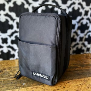 May include: A black rectangular storage case with a top handle and a zippered closure. The front features an open slip pocket and a small rectangular patch with the white text CASE LOGIC stitched onto the lower centre of the fabric exterior.