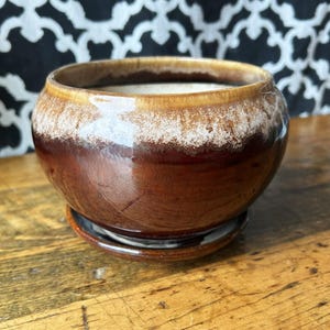 Peut inclure: Un pot de fleurs rond en céramique avec une finition brillante, présentant une base brun foncé qui se transforme en un vernis crème et beige moucheté près du bord. Le pot repose sur une soucoupe circulaire assortie en céramique brun foncé.