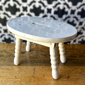May include: A small, oval-shaped wooden stool with a light grey painted top and four cream-coloured, turned legs. The seat features a central rectangular cutout handle and is secured to the base with visible metal screws on the flat surface.