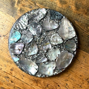 Pode incluir: Base redonda com design de mosaico. A base é feita de resina e incrustada com conchas de abalone iridescentes e pequenas pedras cinza escuro. As conchas têm tons de prata, azul e verde. A base está sobre uma superfície de madeira.