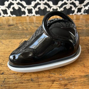May include: A black ceramic butter dish with a glossy finish. The dish has a rounded shape with a handle and sits on a white base. The butter dish is on a wooden surface.
