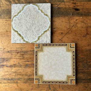 May include: Two square ceramic tiles with decorative designs. One tile has a textured beige background with a white and gold quatrefoil shape. The other tile has a cream-coloured centre with a brown and gold border.