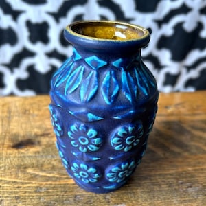 Peut inclure: Un vase en céramique bleu avec un motif floral et de feuilles texturé. Le vase a un extérieur bleu foncé avec des accents turquoise et un intérieur marron. Le vase est posé sur une surface en bois.