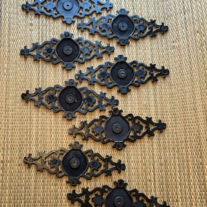 Peut inclure: Six poignées de tiroir décoratives en métal noir avec des motifs ornés et symétriques. Chaque poignée présente un élément central circulaire et des volutes complexes, parfaites pour ajouter une touche vintage aux meubles. Les poignées sont disposées sur une surface texturée brun clair.