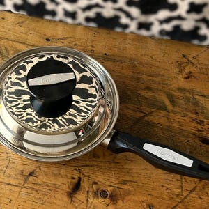 May include: Stainless steel Carico saucepan with a black handle and lid. The lid has a black and white animal print design and a black knob. The handle has a white Carico label.