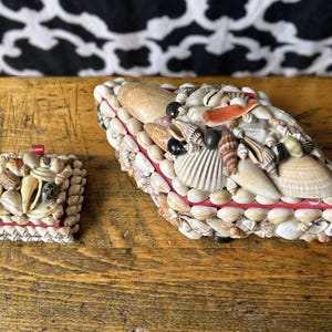May include: Two boxes decorated with seashells. The larger box is oval, the smaller is square. Both have red trim and are covered in various shells. They sit on a wooden surface. The background is black and white.