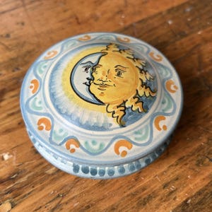 May include: A round ceramic trinket box featuring a hand-painted design of a yellow sun and a blue crescent moon on the lid. The border is decorated with a repeating wavy pattern in light blue, white, and orange, with small blue dots along the base.