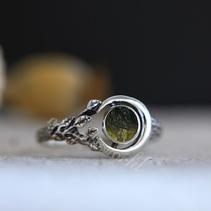 Peut inclure: Bague en argent ornée d'un croissant de lune sertissant une pierre ronde vert olive. La monture de la bague présente un motif détaillé inspiré de la nature, avec des feuilles et des branches, créant un bijou unique et élégant.